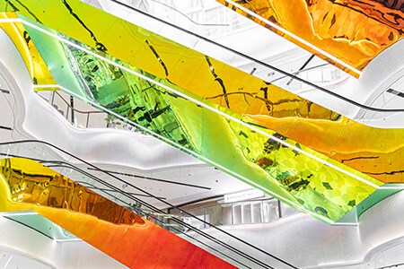 Winkelcentrum met meerdere verdiepingen, witte wanden en hangende roltrappen bedekt met kleurrijke, reflecterende 3M dichroïsche glasafwerkingen met rode, gele, oranje en groene tinten.
