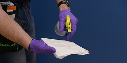 Iemand met blauwe wegwerphandschoenen die een keukenrol besproeit met de verbeterde 3M hechtingsspray
