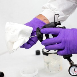 Handen met paarse handschoenen die een spuitpistoolnaald schoonmaken met een wit doekje