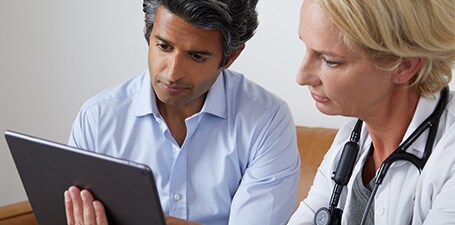 Behandelaar ausculteert en toont app op tablet aan man
