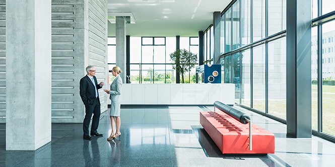 Panoramisch beeld van een lobby in een kantoorgebouw omgeven door glazen wanden, met twee zakenmensen in gesprek
