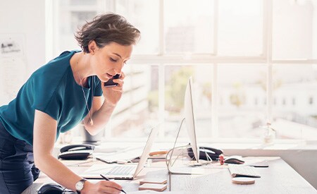 Vrouw met telefoon die aantekeningen maakt in een helder, zonnig kantoor met grote ramen
